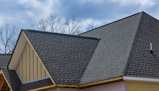 A low-angle view of a grey shingled roof on a house under construction against a cloudy blue sky.