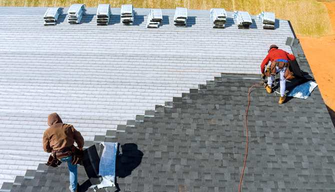 Two people in work gear install dark asphalt shingles on a roof, transitioning from an underlayment-covered surface.