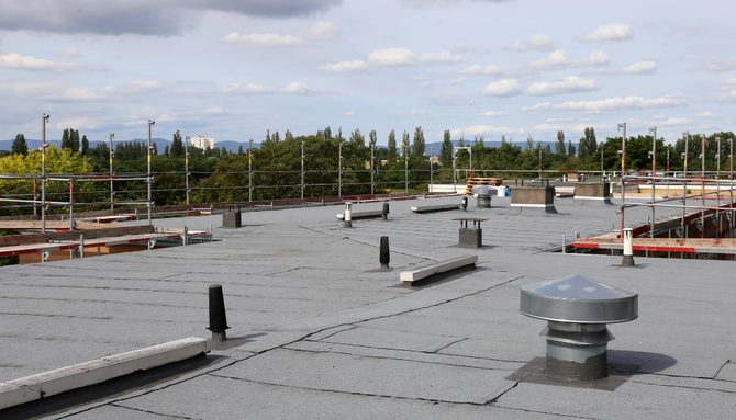 A grey commercial flat roof with various vents, railings, and pipes under a cloudy blue sky.