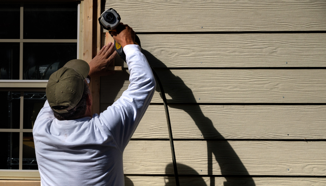 A person wearing a white long-sleeve shirt and cap uses a nail gun to install wood siding near a window.