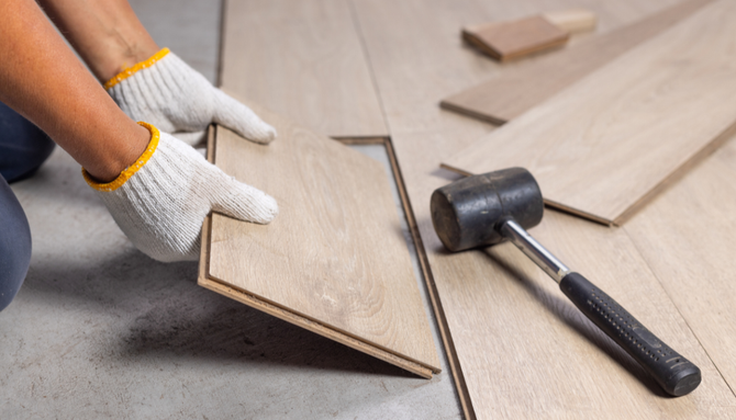 Hands wearing white work gloves installing light-colored wood laminate flooring with a rubber mallet nearby.