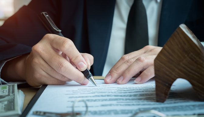A person in a suit signs a real estate contract next to a miniature wooden house model and a stack of cash.