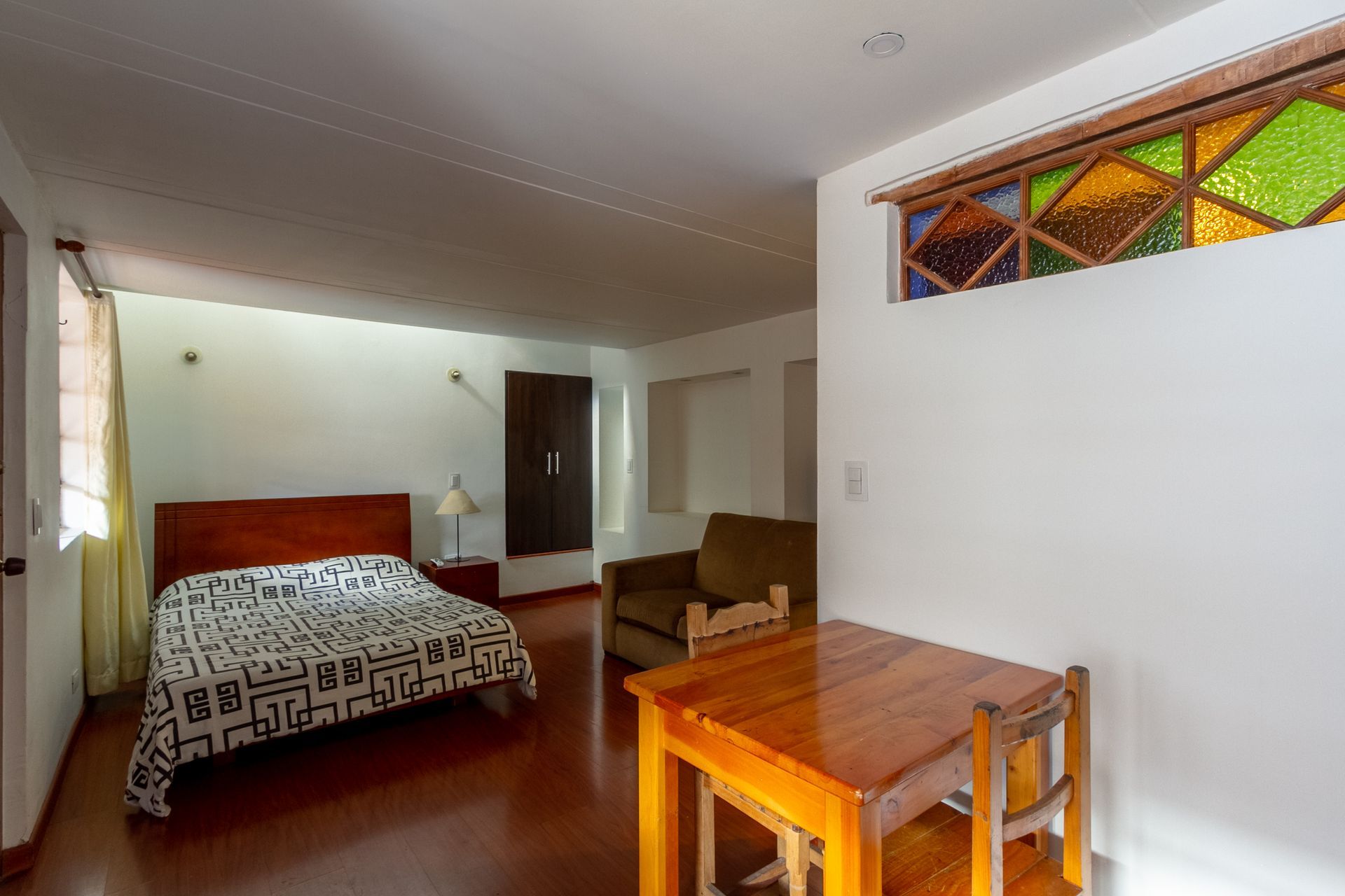 A bedroom with a bed , table , chairs and a stained glass window.