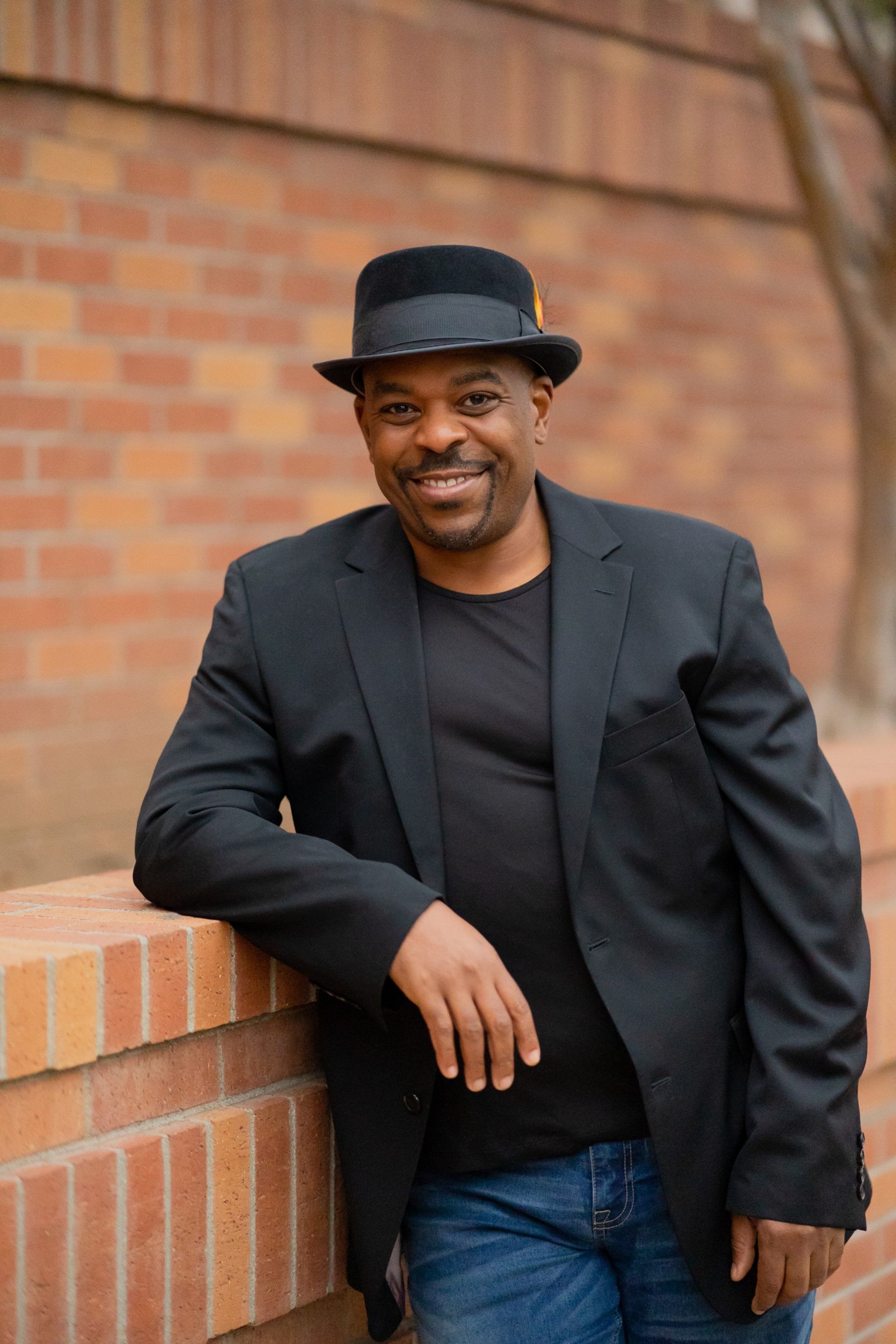 A man in a suit and hat is leaning against a brick wall.