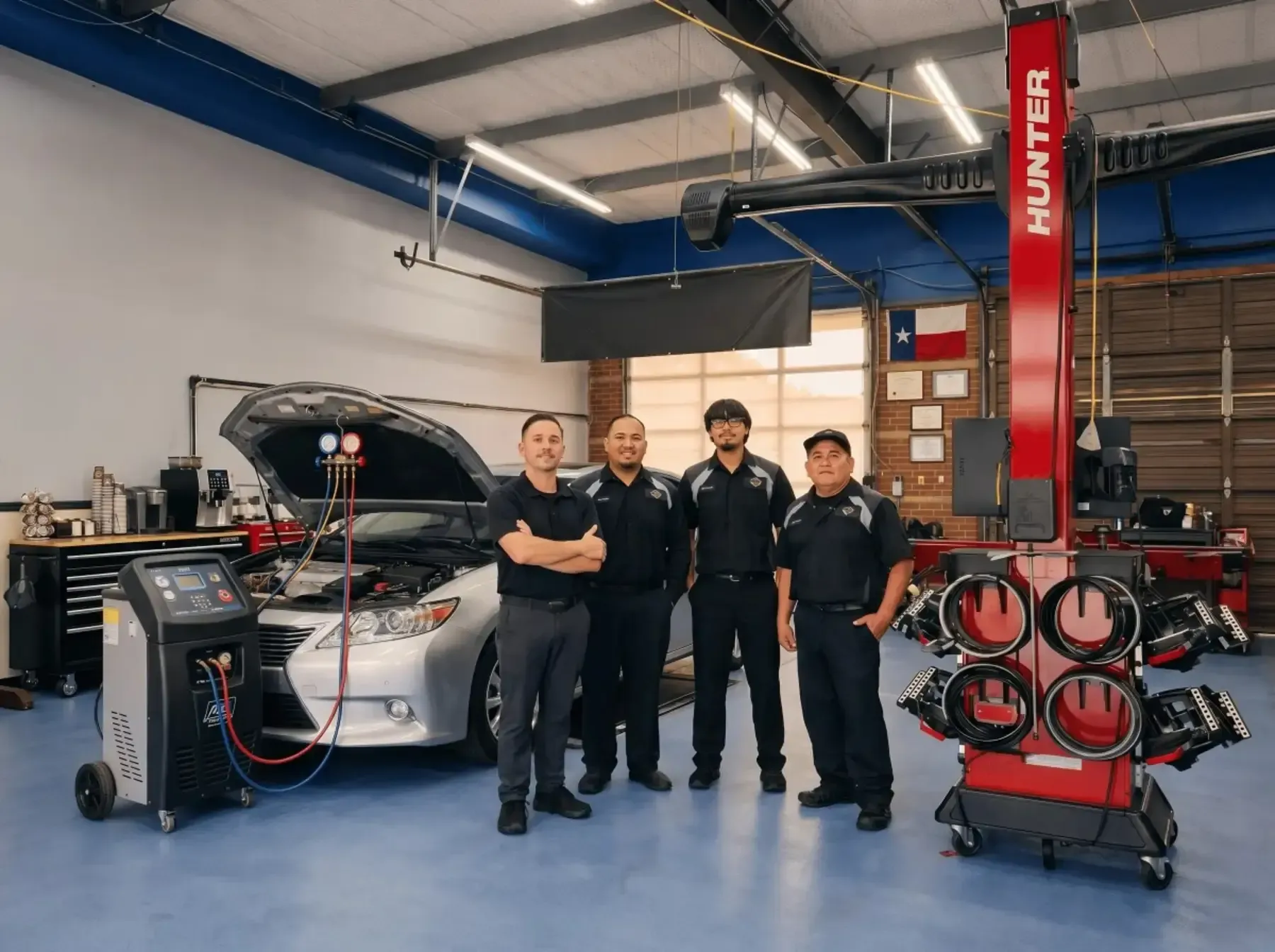 Four mechanics standing in an auto shop beside a silver car with its hood open and a tire machine nearby