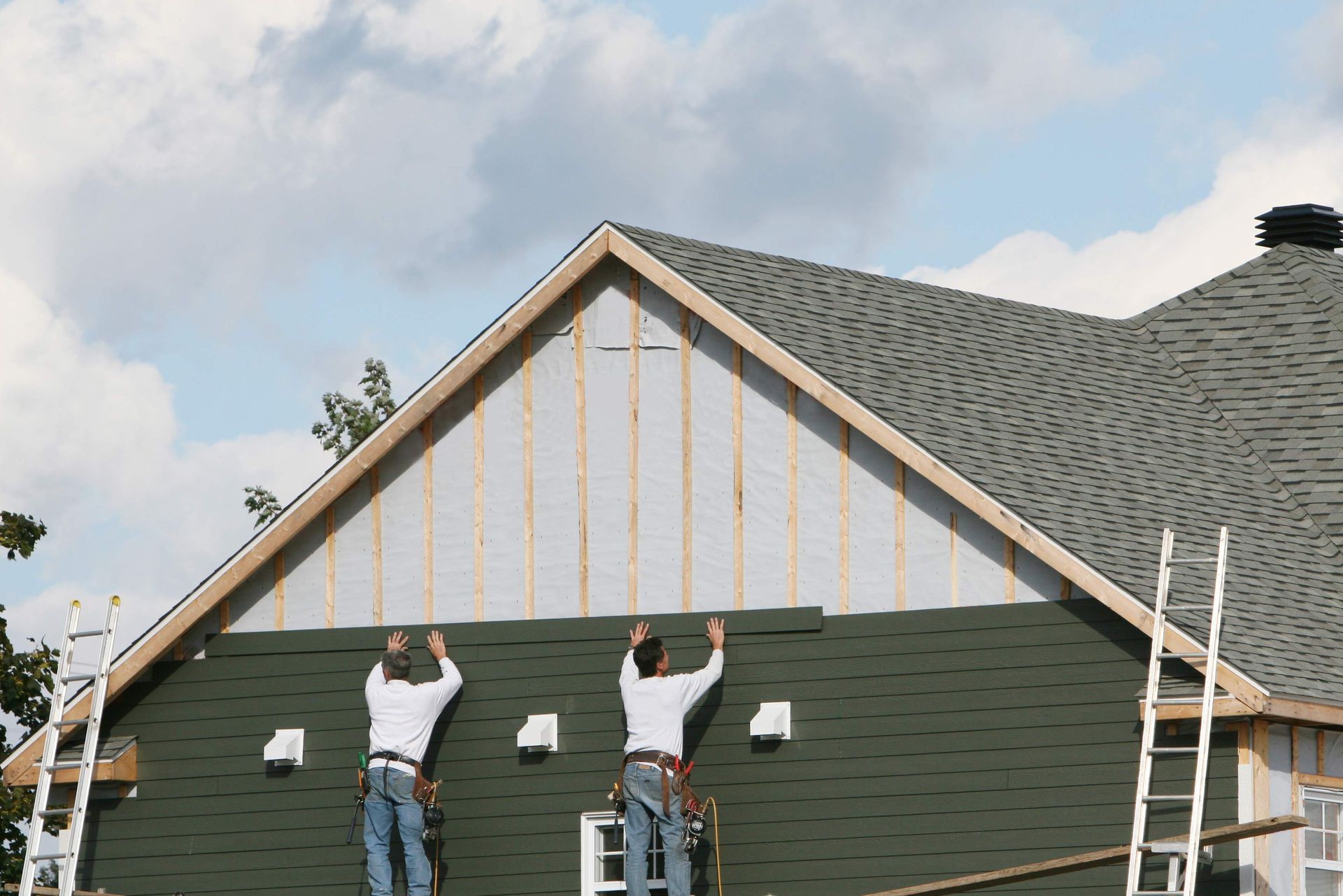Two people installing green shingles on a house's gable under a cloudy sky.