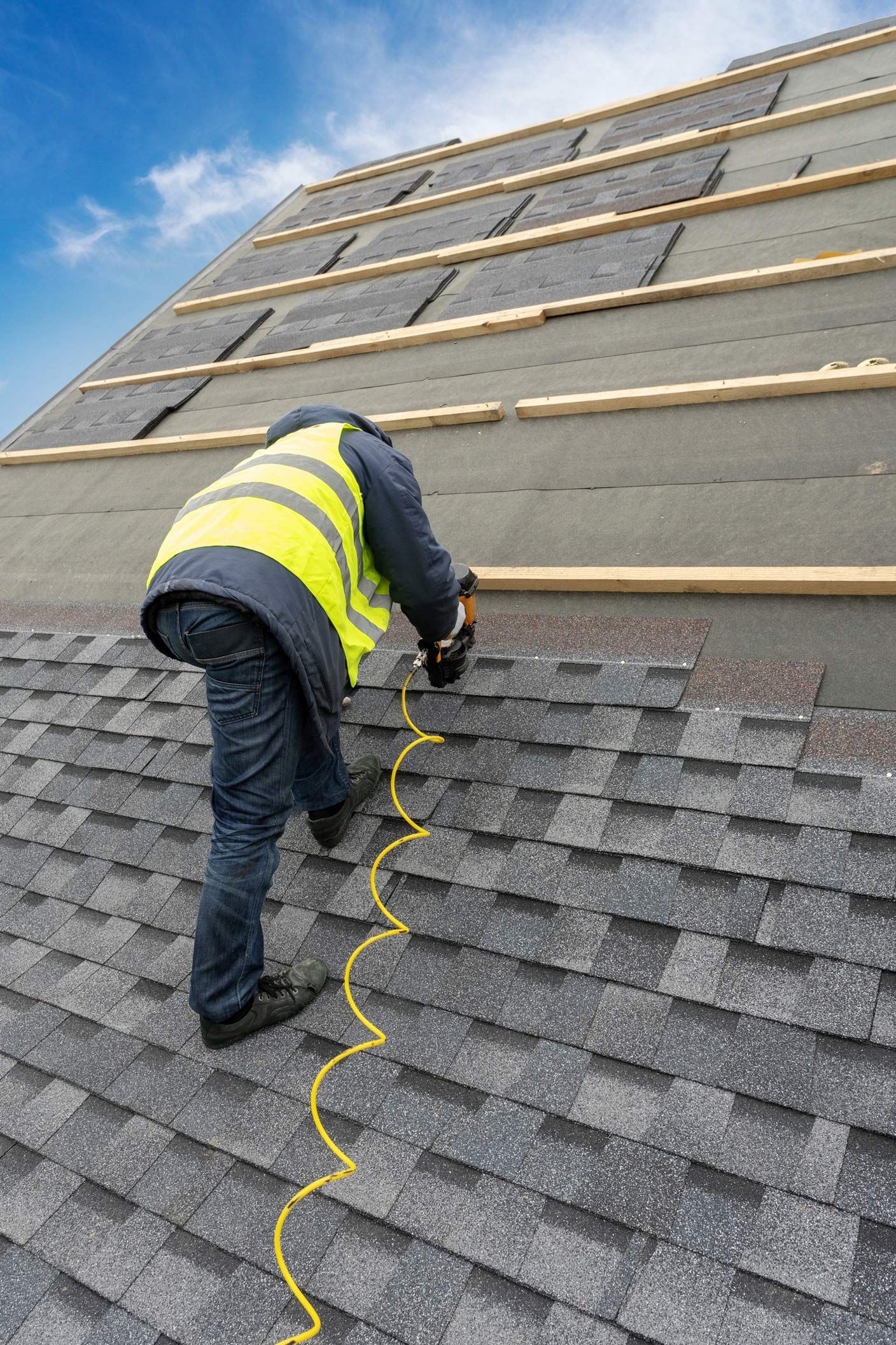 Roofer in safety vest using a nail gun on a shingled roof under a bright sky.