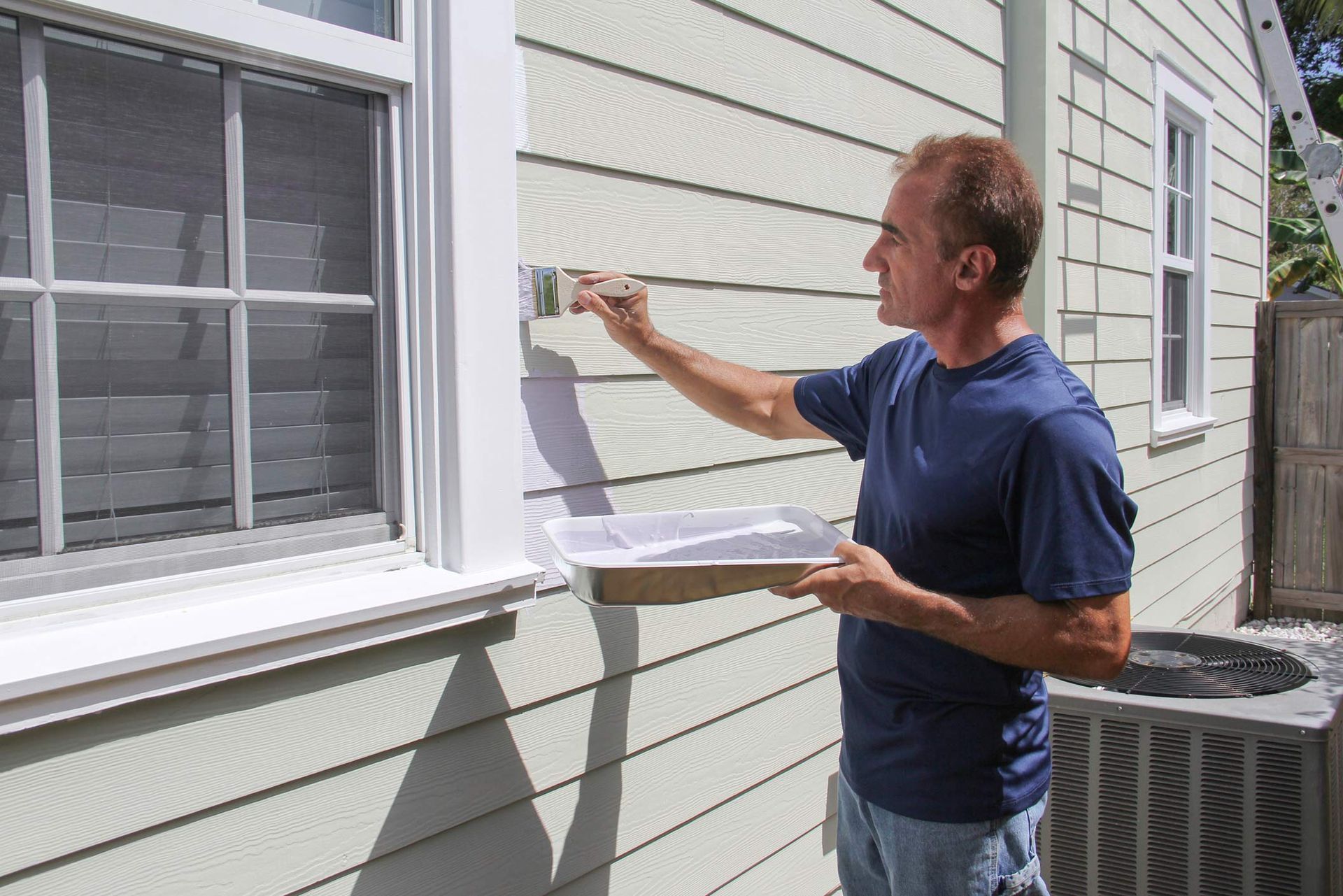 Man paints house siding with a paintbrush, holding paint tray. Exterior of a house with window.