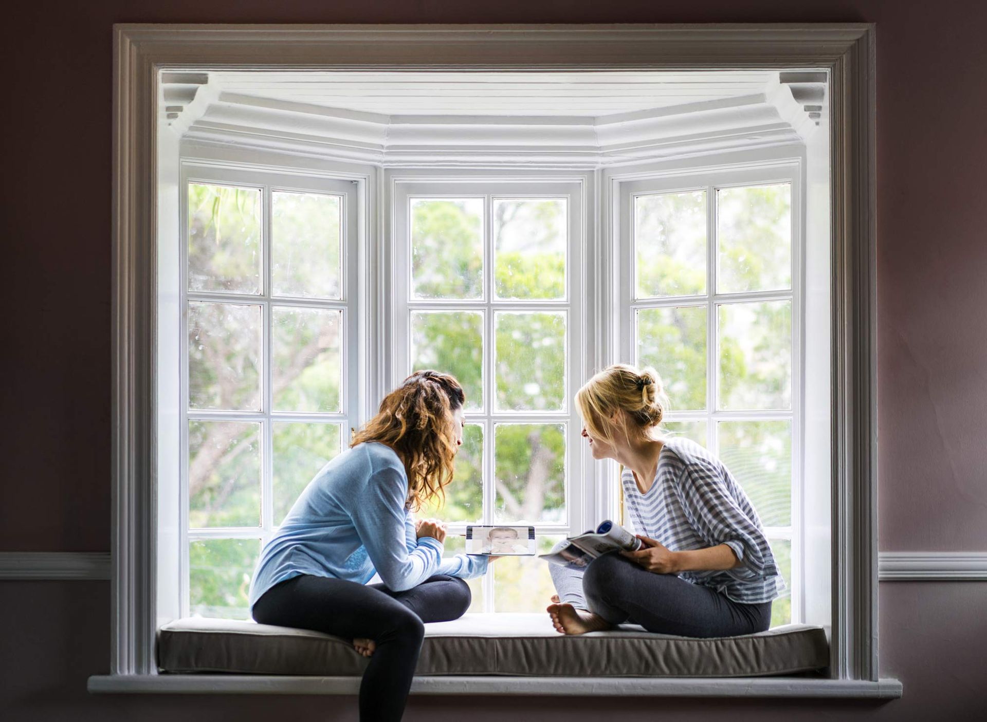 Two people sit in a bay window, looking at something together. Sunlight streams through the window.