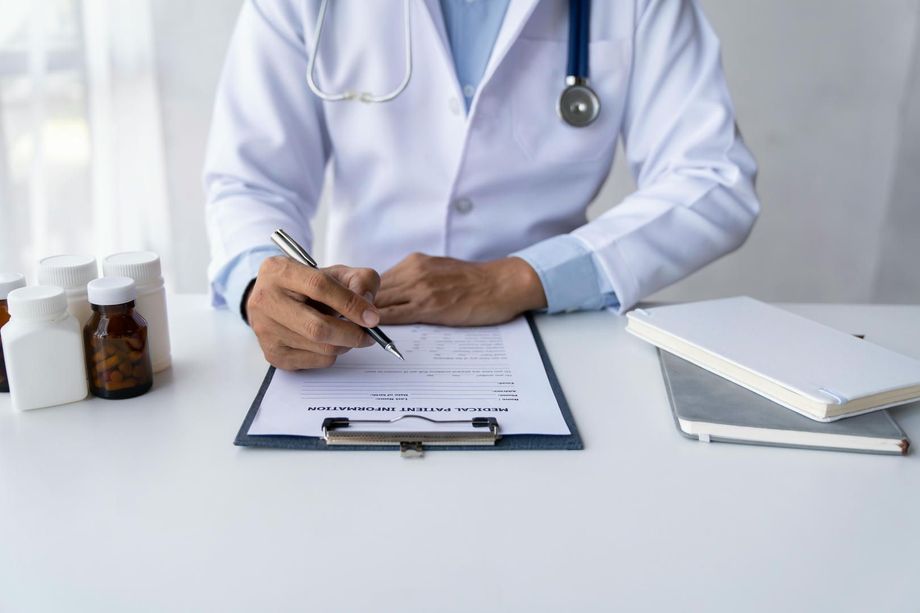 Doctor in white coat writing on a clipboard at a desk with medicine bottles.