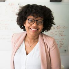 Woman with glasses, curly hair, smiling, wearing a pink blazer in front of a whiteboard.