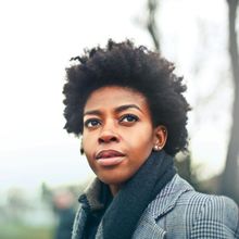Black woman with natural hair, looking up, wearing a patterned coat and scarf, outdoors.
