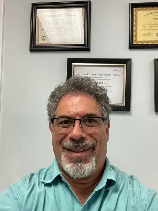 Middle-aged man with glasses and a beard smiling, wearing a teal shirt; framed certificates on a white wall behind him.