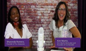 Two women are sitting at a table with a microphone in front of a brick wall.