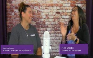 Two women are sitting at a table with a microphone in front of a brick wall.