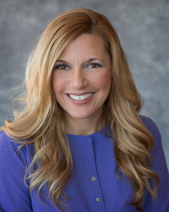 A woman with long blonde hair is wearing a purple sweater and smiling.