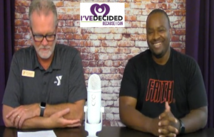 Two men are sitting at a table with a microphone and a sign that says i ve decided