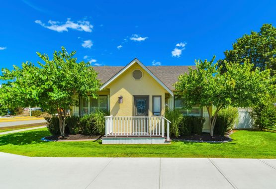 A small yellow house with a porch and trees in front of it.