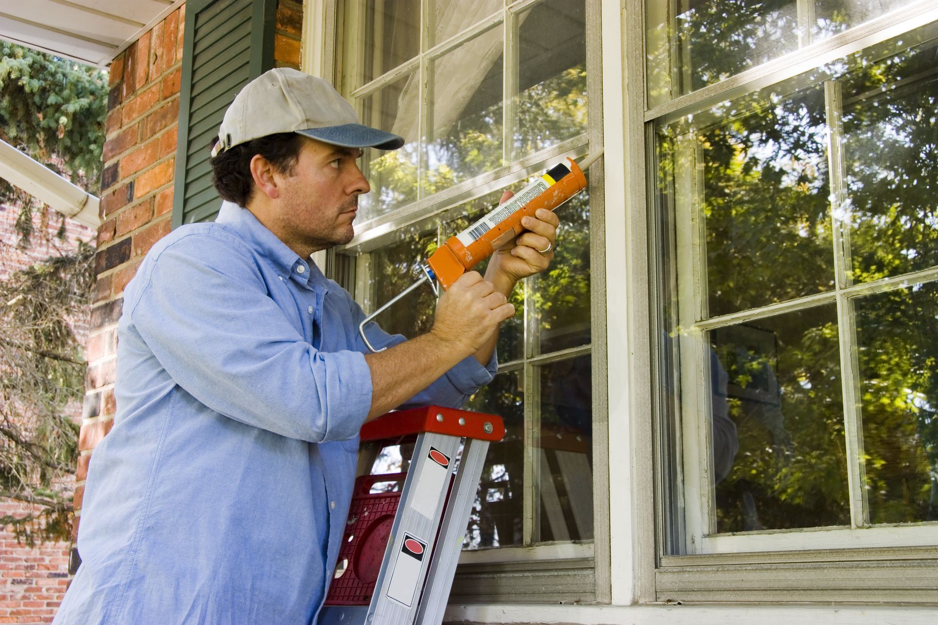 A man is standing on a ladder applying caulk to a window.