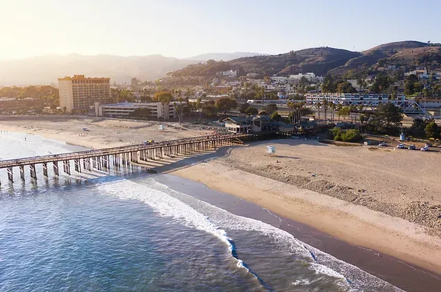 An aerial view of a beach with a pier and mountains in the background.