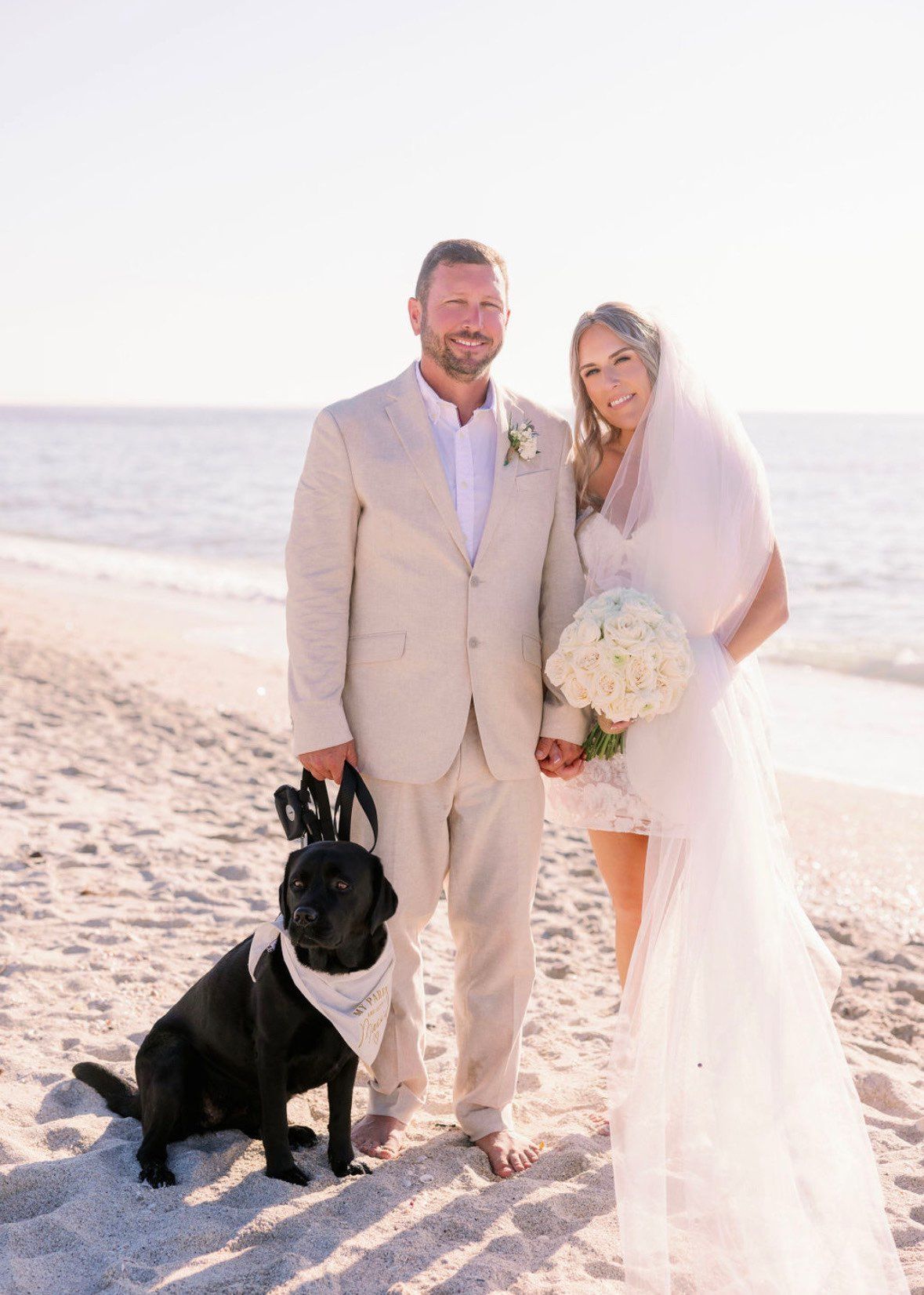 Bride and groom with dog on beach; dog wears bandana and bunny ears, holding hands. Ocean in background.