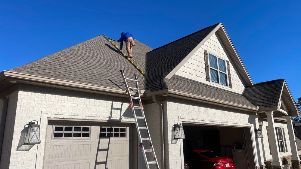 Person on a ladder measuring a brown shingled roof of a house with a white exterior and two garage doors.