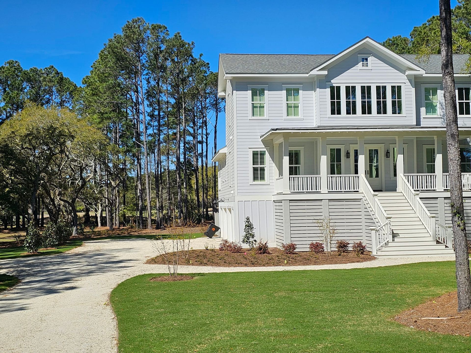 Light gray two-story house with porch, white steps, and gravel driveway on green lawn, surrounded by trees.