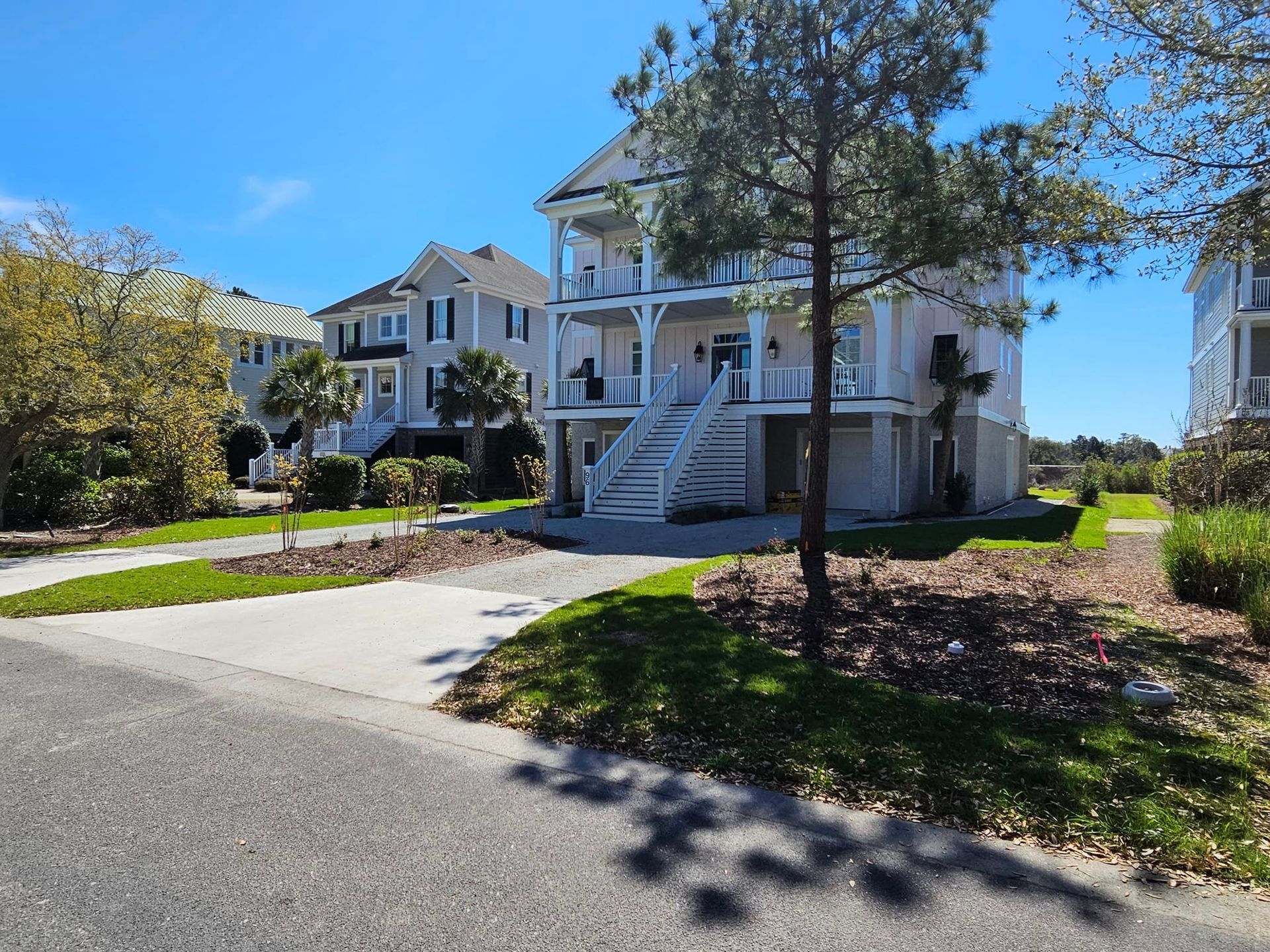Two-story house with white exterior, balconies, and stairs leading to the front door, set on a paved road with grass and trees.