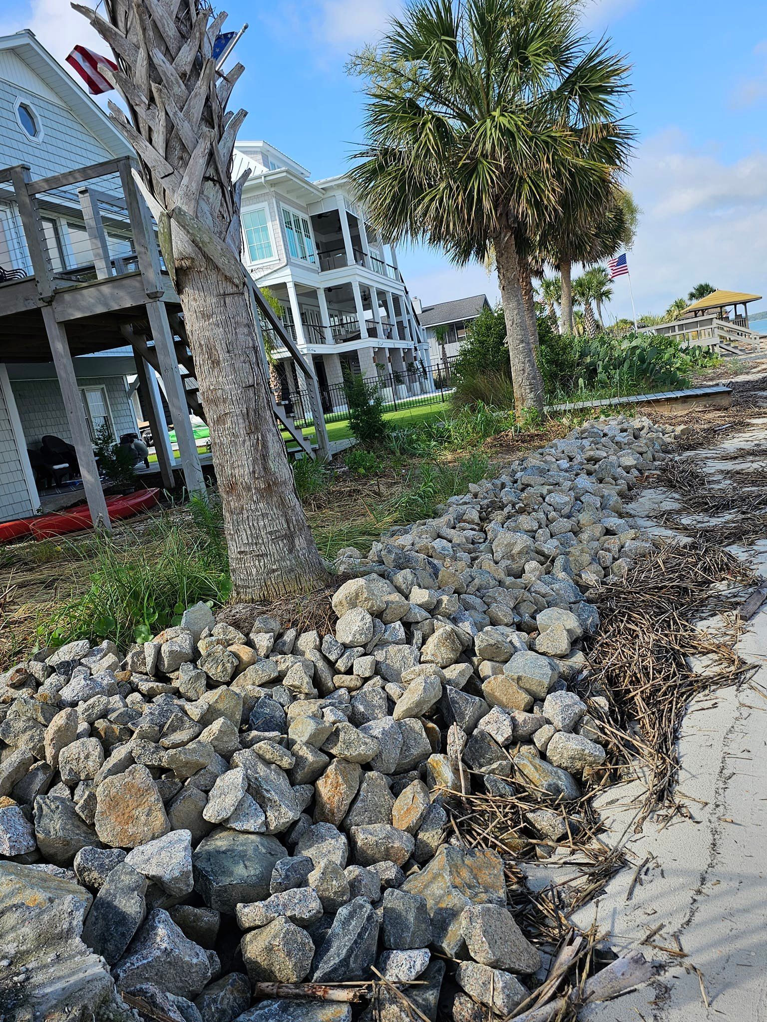Coastal view: Rock barrier in front of beach houses, palm trees and blue sky.