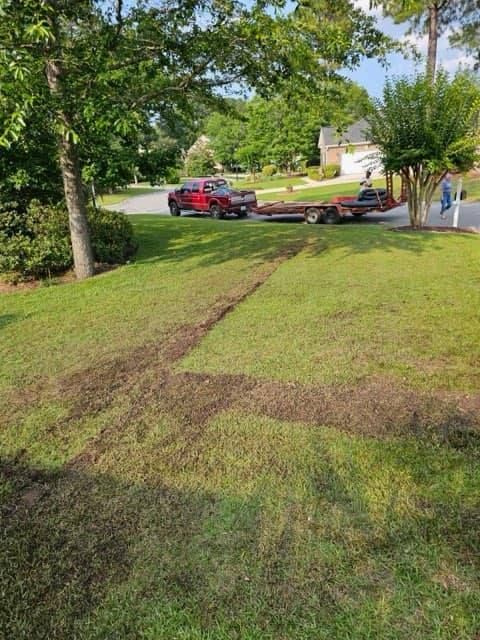 A red truck with a trailer on a lawn has left tire tracks. Green grass, trees, and a house in the background.