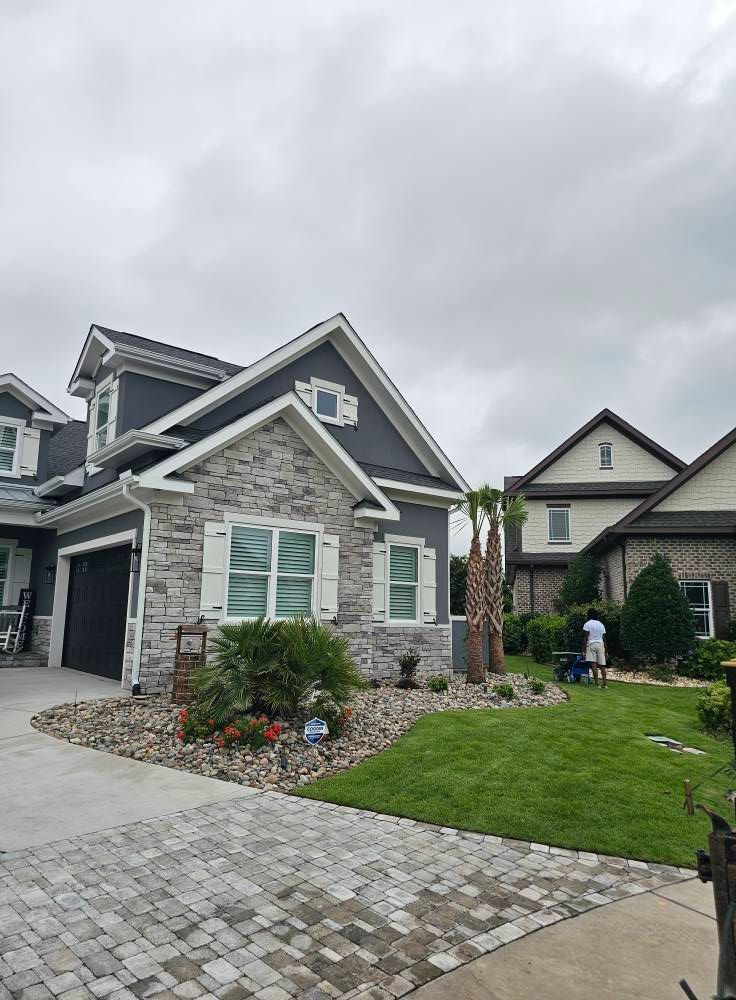 House with gray siding, brick facade, and a cobblestone driveway under a cloudy sky.