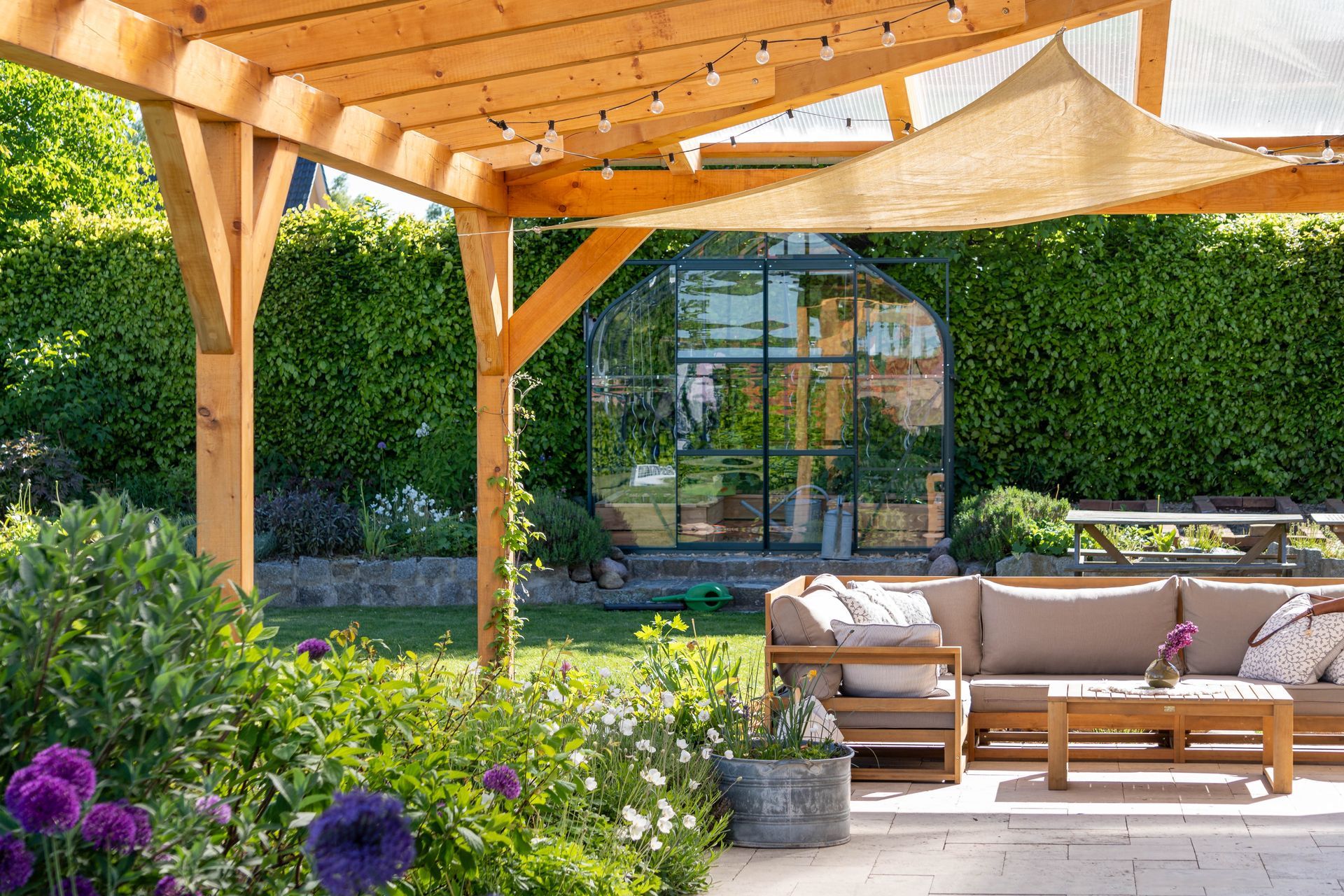 Wooden pergola over a patio with outdoor furniture; greenhouse and garden backdrop.