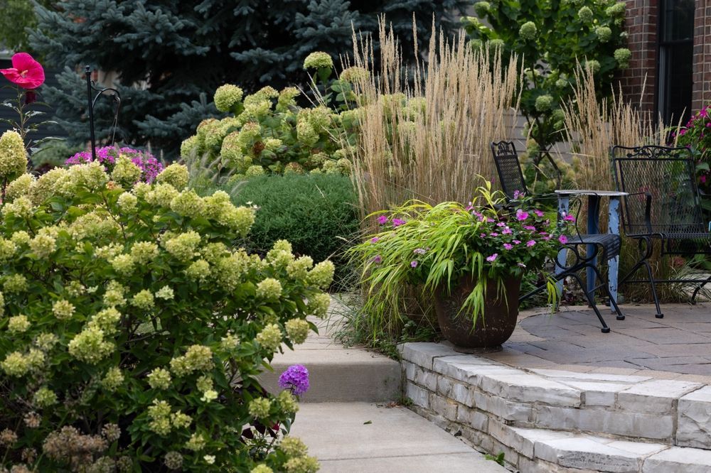 Lush garden with steps, stone patio, and patio furniture, featuring green, beige, and pink flowers.