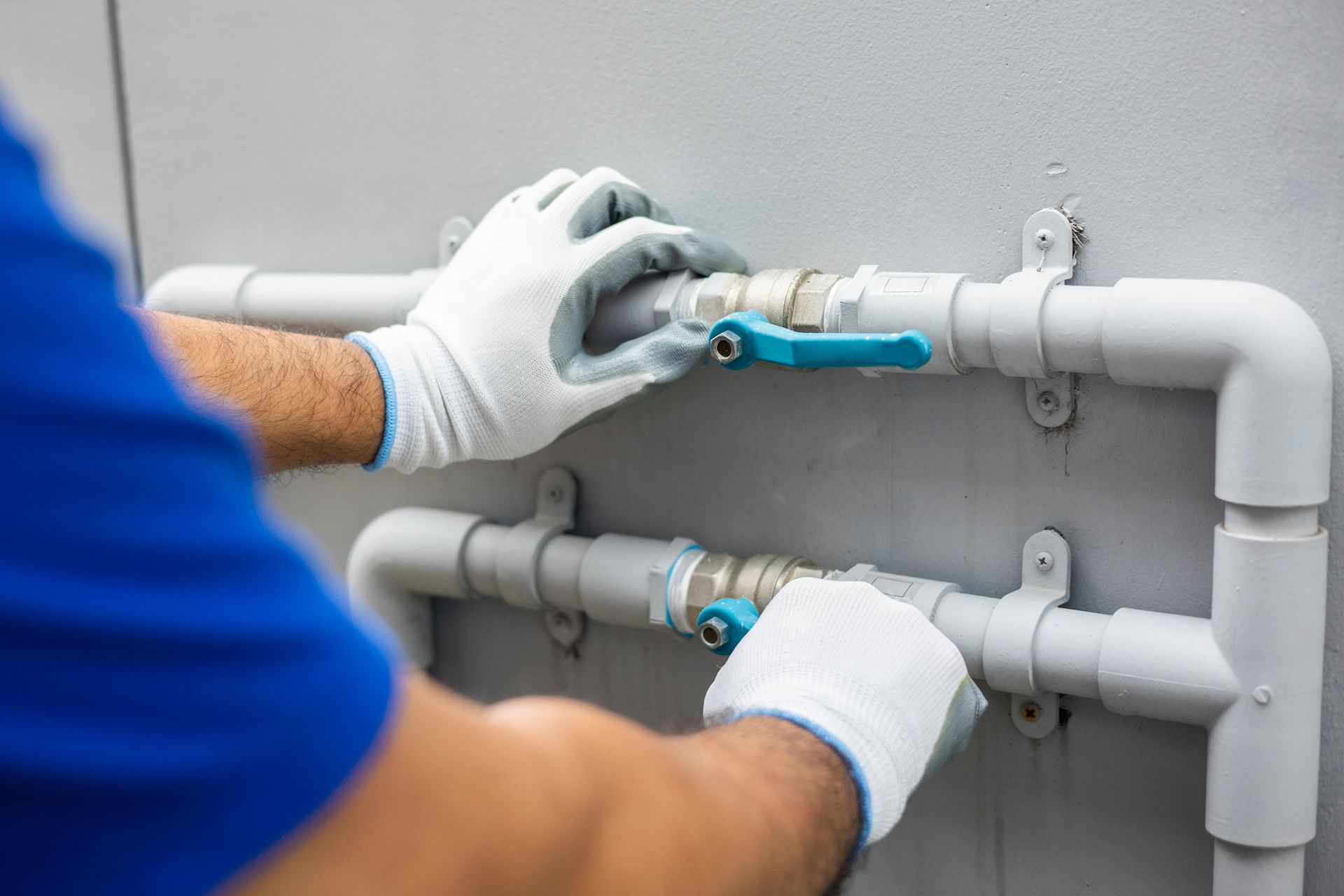 Gloved plumber tightening a blue valve on white PVC pipes against a gray wall