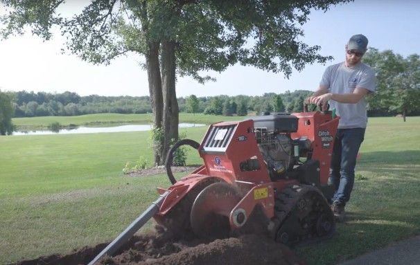 Man operating a red trencher in a grassy area, digging a trench near a path and a body of water.