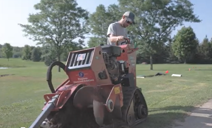 A person operates a red tracked trencher machine on a grass field under a sunny sky.
