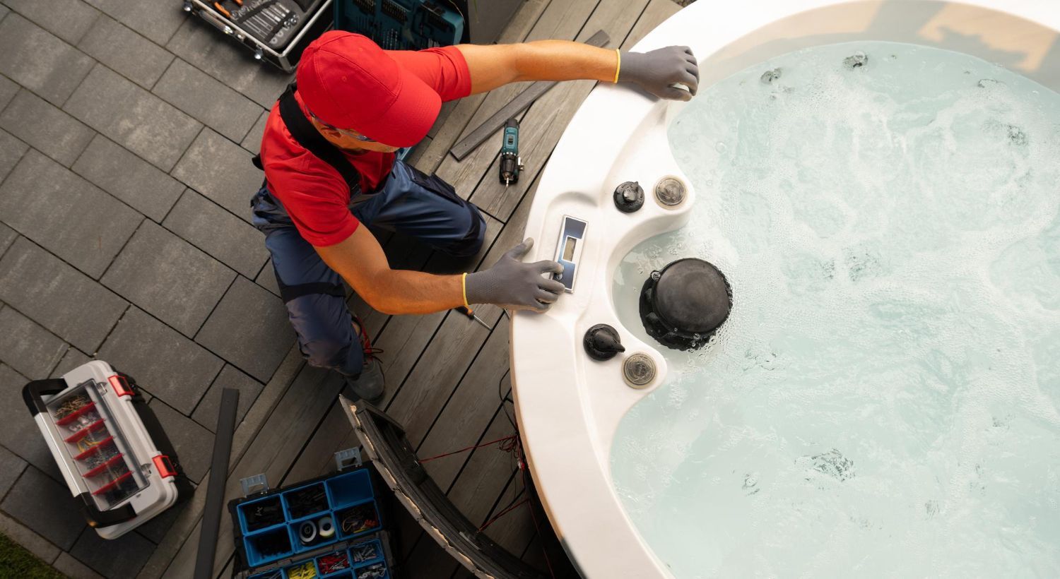 A technician in a red shirt and work pants repairs a hot tub on a paved patio, surrounded by tool kits.
