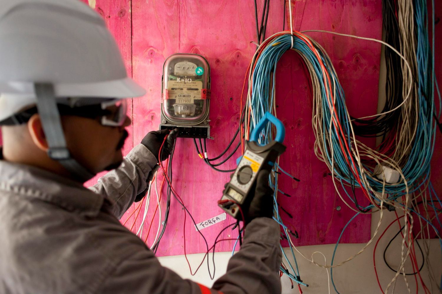 Electrician in safety gear working on wires near a meter. Pink wall and colorful wires.
