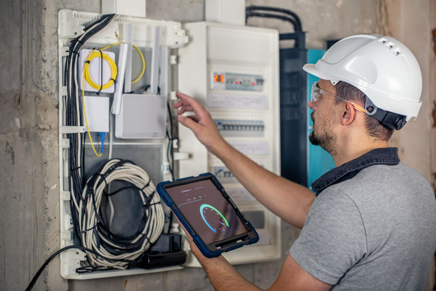 A man in a hard hat is using a tablet to look at an electrical box.