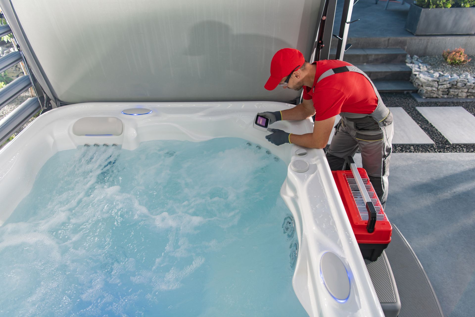 A person in red shirt and cap repairs a hot tub outdoors, toolbox nearby.