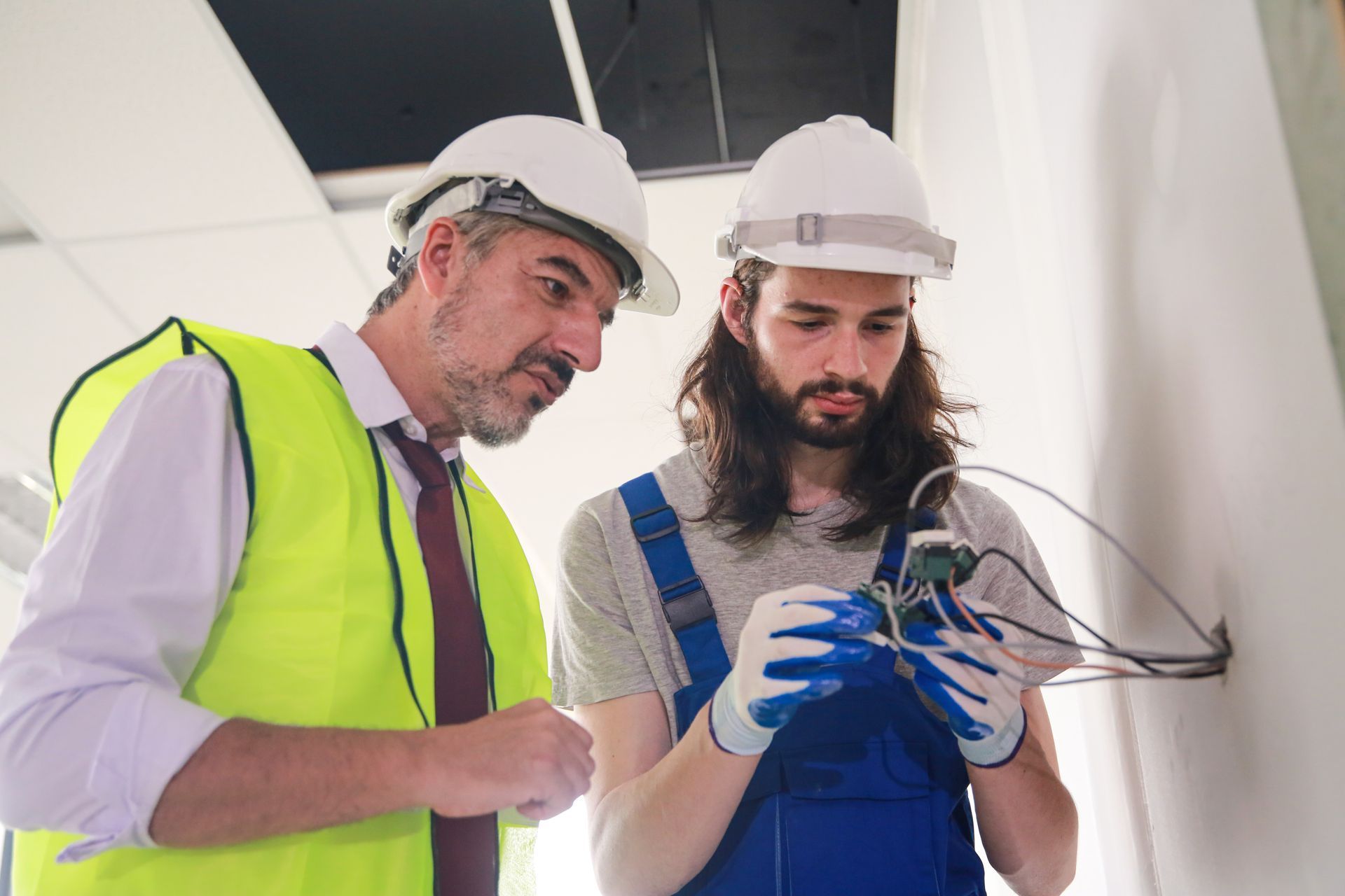 A man in a hard hat is standing next to a man in overalls.