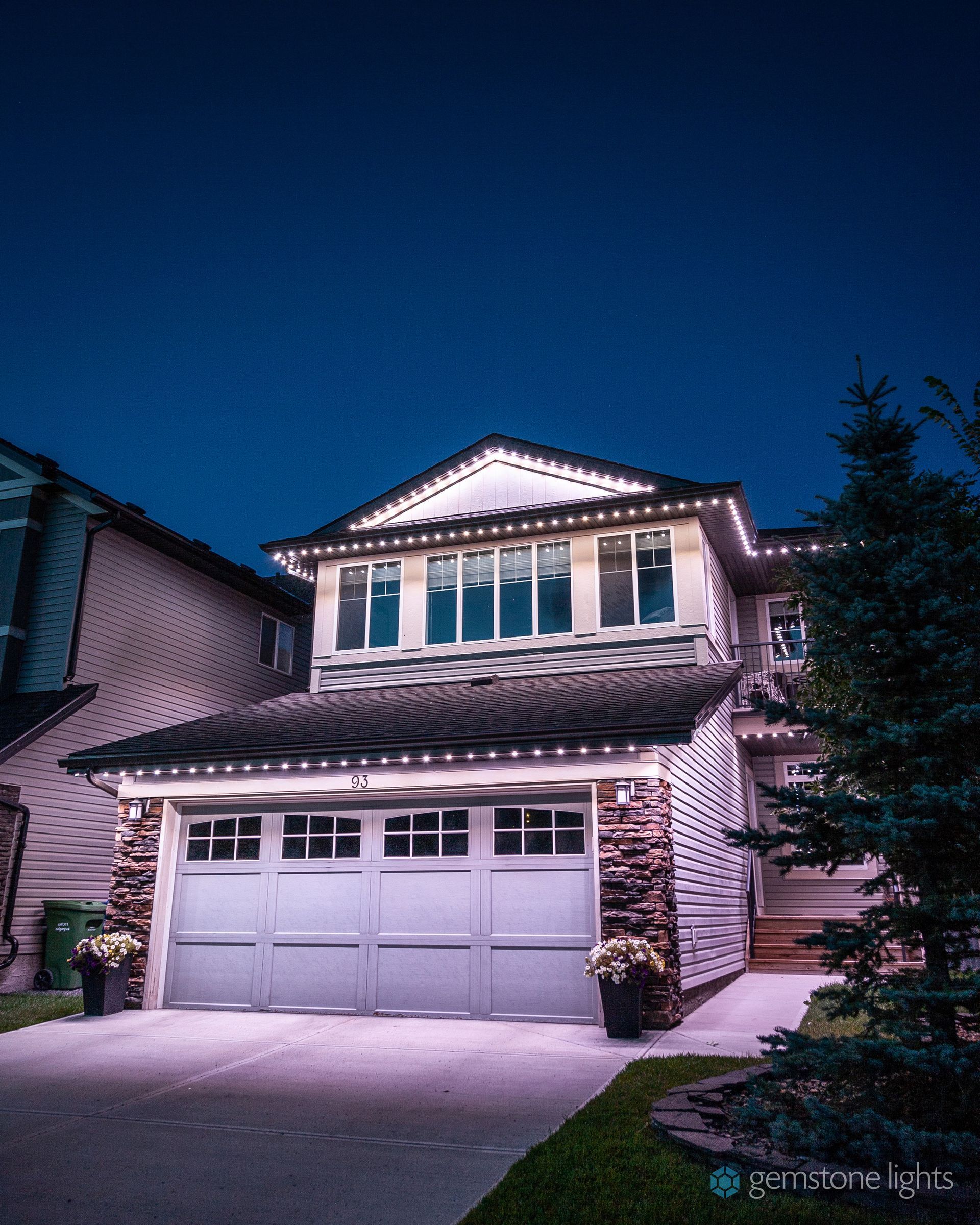Two-story house with Christmas lights at night. White garage door, dark roof, blue sky, and a few evergreen trees.