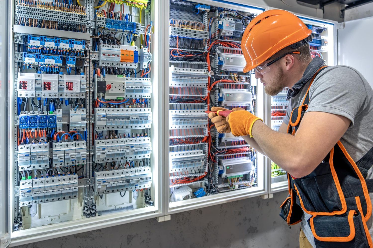 Electrician working on a complex electrical panel, wearing safety gear, in a utility room.