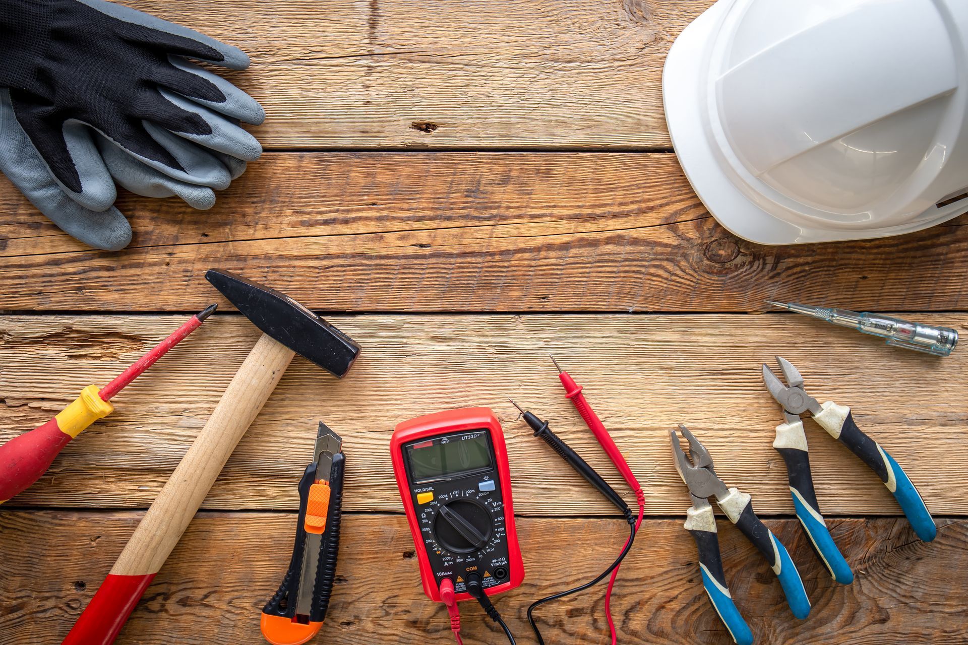 A hard hat , gloves , tools , and a multimeter are on a wooden table.