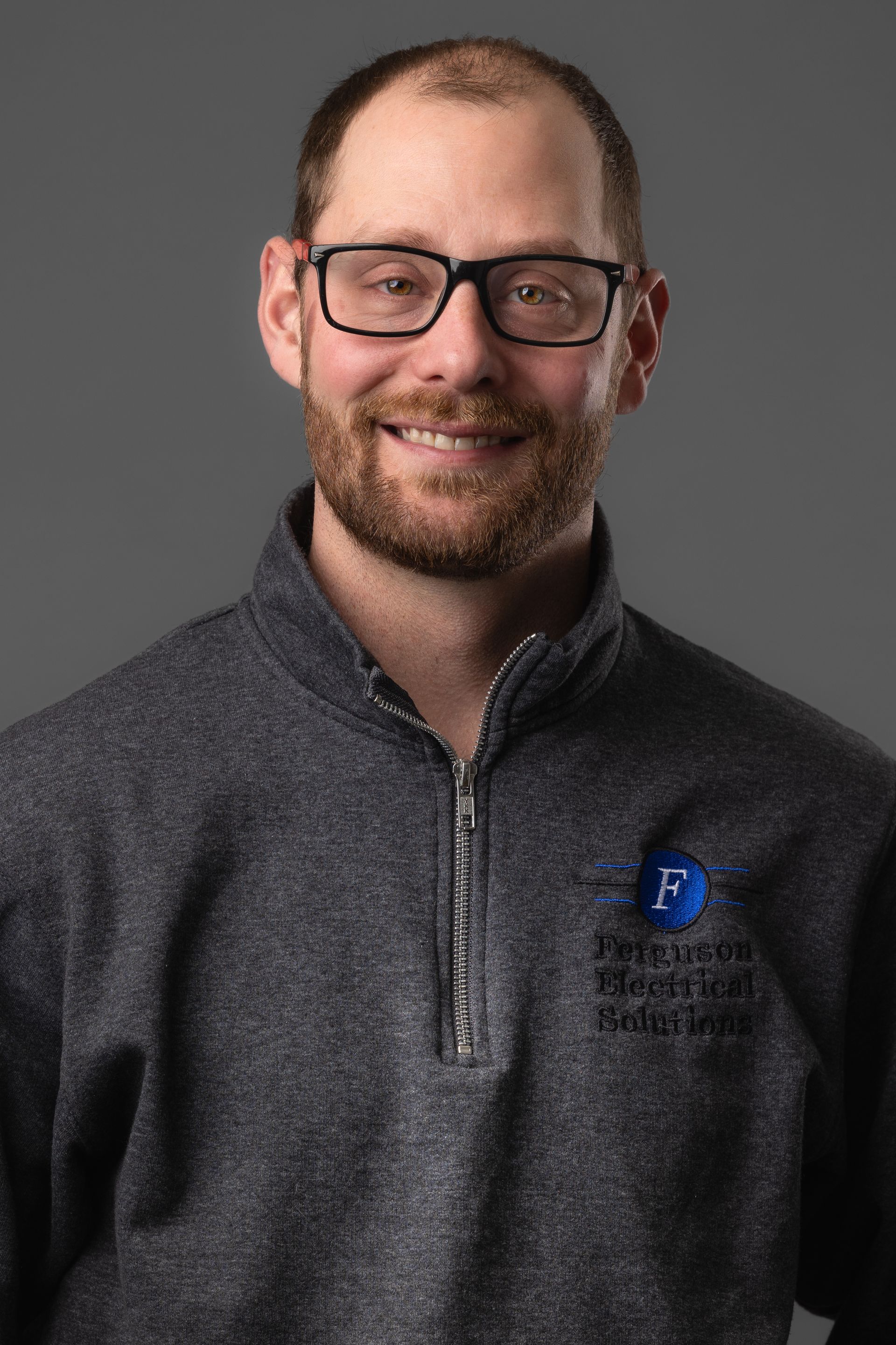 A man with a beard and glasses is wearing a gray sweater and smiling.