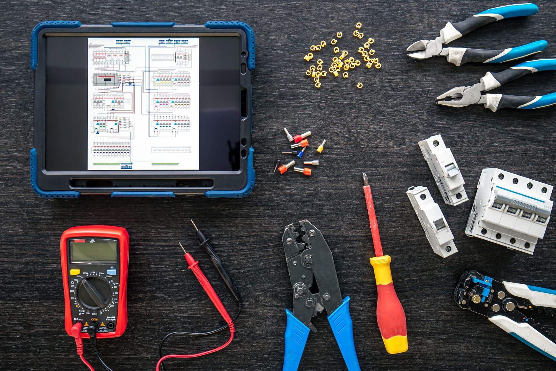 An electrician's workspace with a tablet showing a wiring diagram, surrounded by tools like pliers, a multimeter, and circuit breakers.