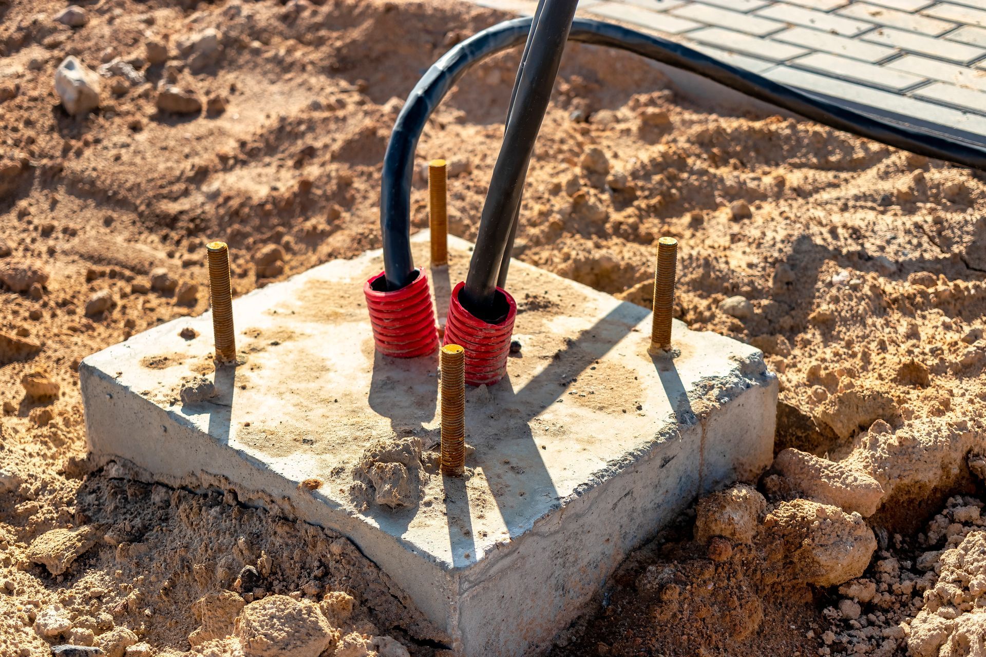 Concrete base with electrical cables and rebar stakes in dirt. Cables are protected by red sleeves.