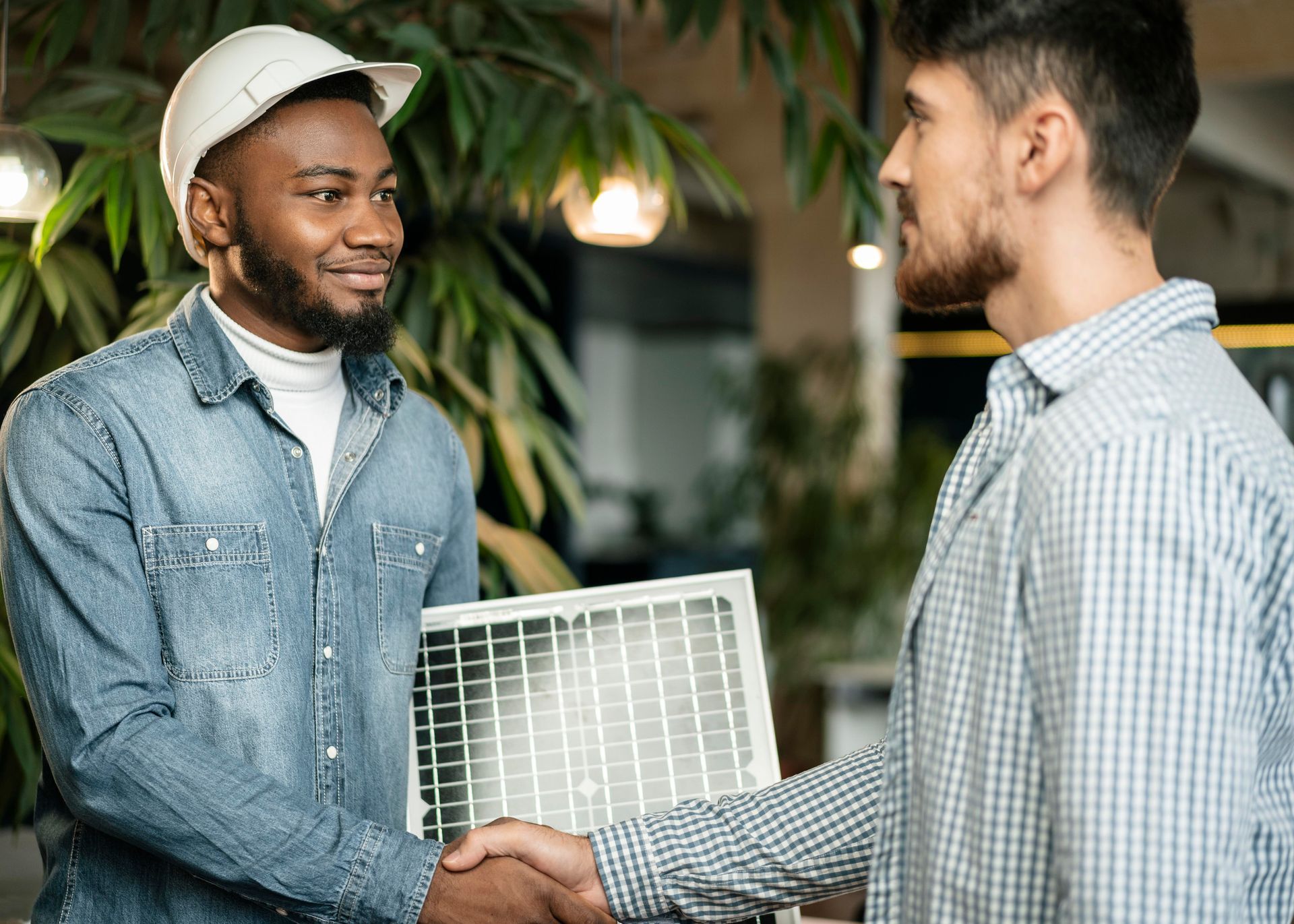 Two men shake hands, one wearing a hard hat and holding a solar panel, likely after a business agreement in an office setting.