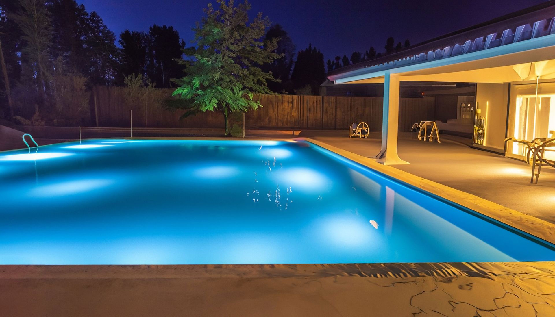 Lit-up pool at night next to a house with a patio. Lights illuminate the blue water.