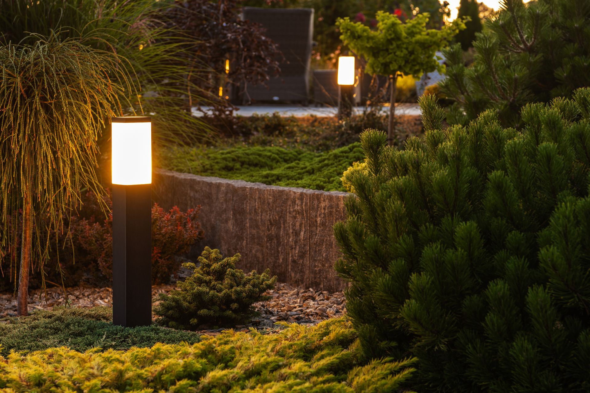 Illuminated garden with modern black pillar lights among various shrubs and plants, at dusk.